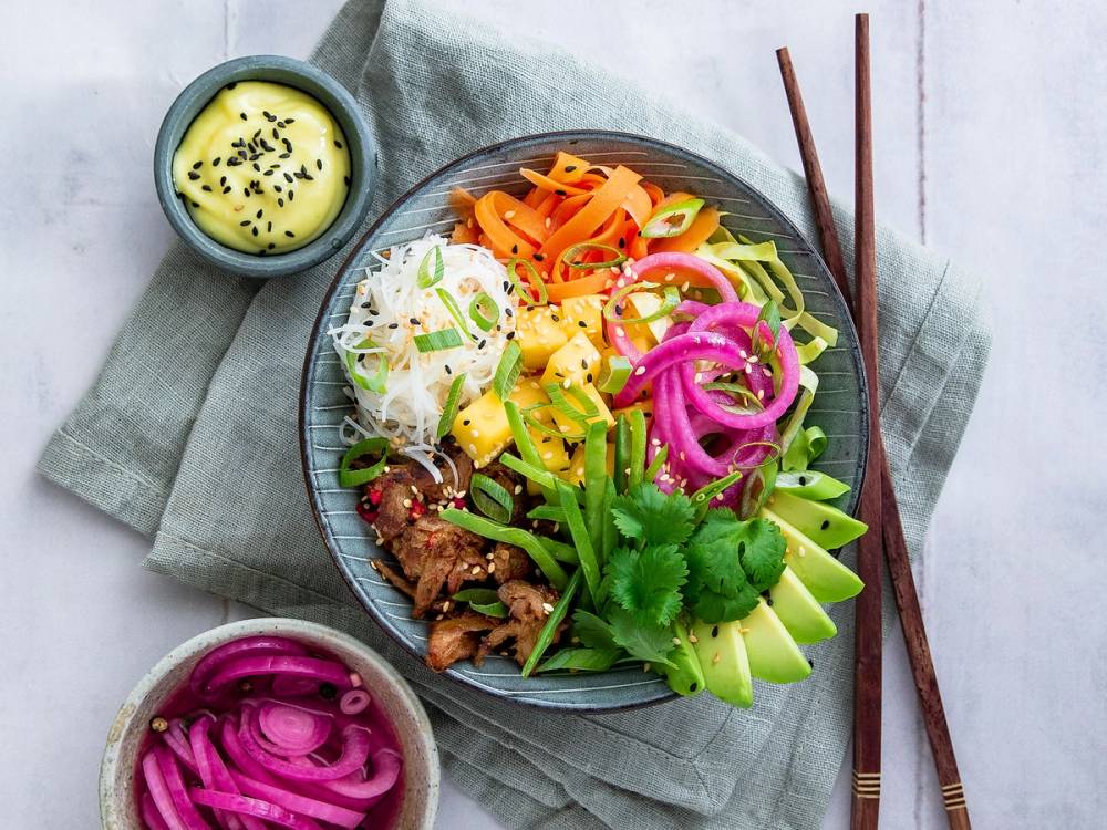 En fargerik vegetarisk lunsjbowl med grønnsaker, avokado, og en liten skål med saus ved siden av, på et bord med spisepinner.