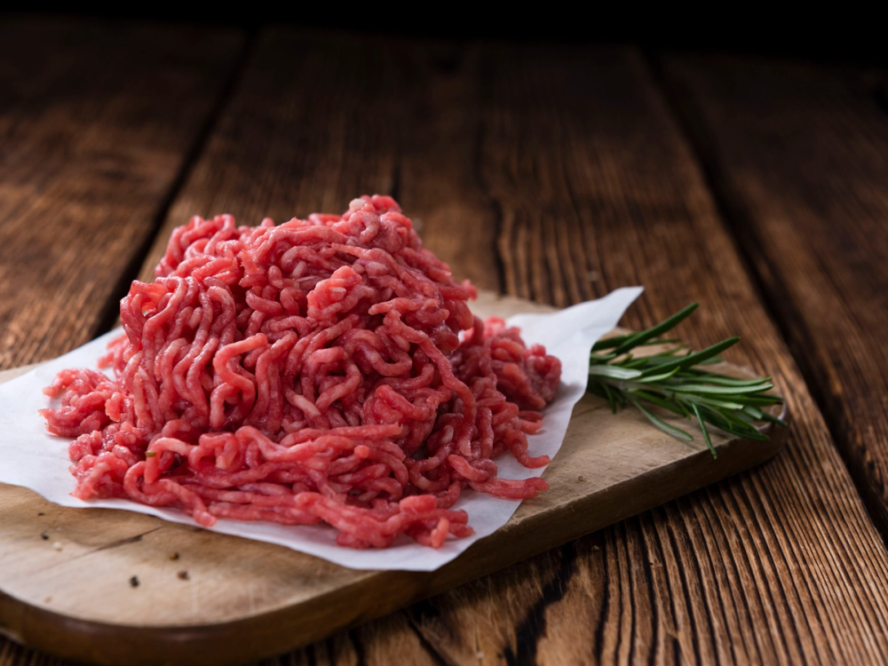 Kjøttdeig på en skjærefjøl med noen grønne urter ved siden av, på et trebord.