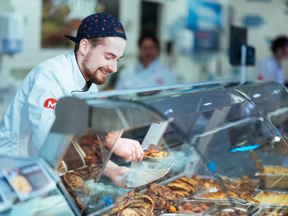 En ansatt i en varmeskapsbutikk med varm mat bak glass, som serverer mat til kunder.