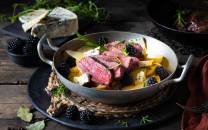 Stekt biff med svartbær og sitron i en hvit serveringsskål på et bord med urter og tilbehør.