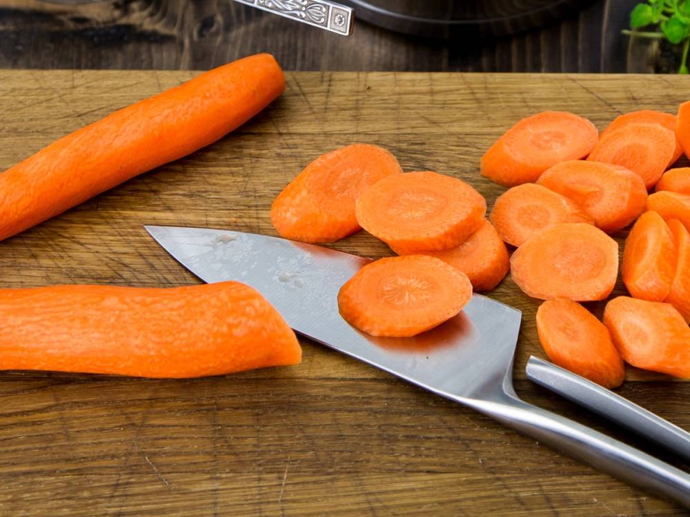 Skiver av gulrot og en hel gulrot på et skjærebrett med en kniv, klar til å kuttes opp.