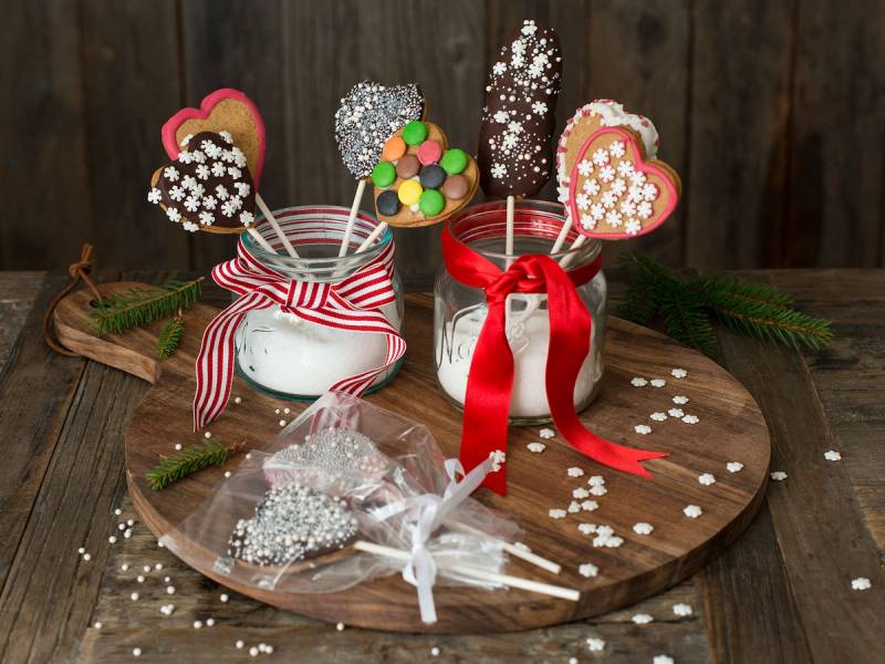 To glass med pepperkaker på pinne pyntet med fargerike og hvite glasurdekorasjoner, omgitt av små pepperkaker og granbar på et rundt trefat, med en trevegg i bakgrunnen.