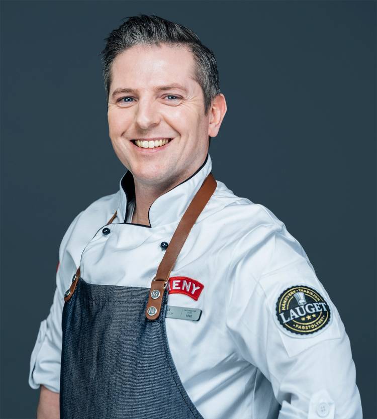 Kokk i hvit uniform og blå forkle med logo, med ansiktet dekket av en grå blokk, står mot en mørk bakgrunn.