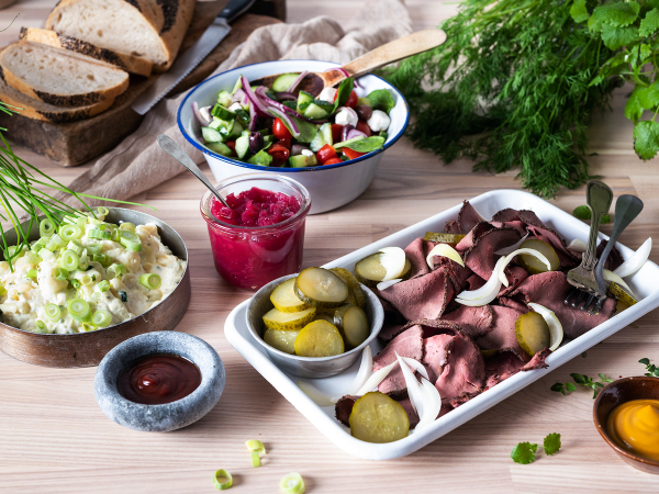 Koldtbord med ulike pålegg, grønnsaker og tilbehør på et bord, inkludert en skål med syltede agurker og en bolle med rød saus.