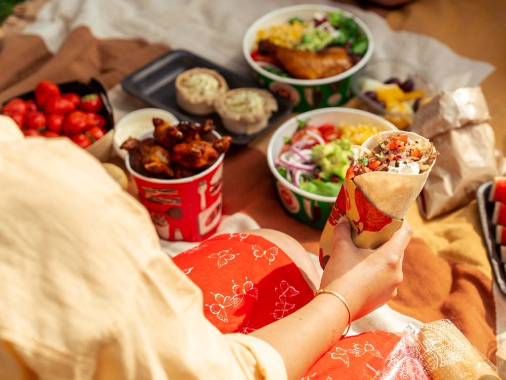 Person som holder en burrito med salat, ost og kjøtt, omgitt av ulike retter og snacks på et bord.