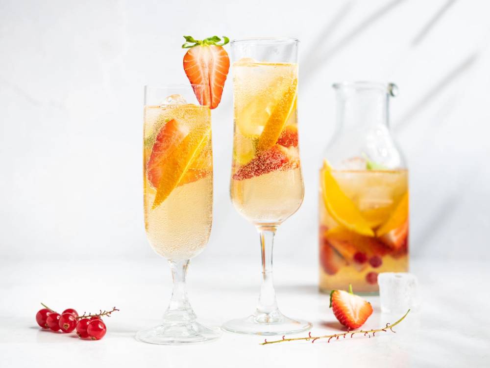 To glass med sommerpunch med frukt og en liten jordbær på kanten, og en mugge med mer sommerpunch i bakgrunnen