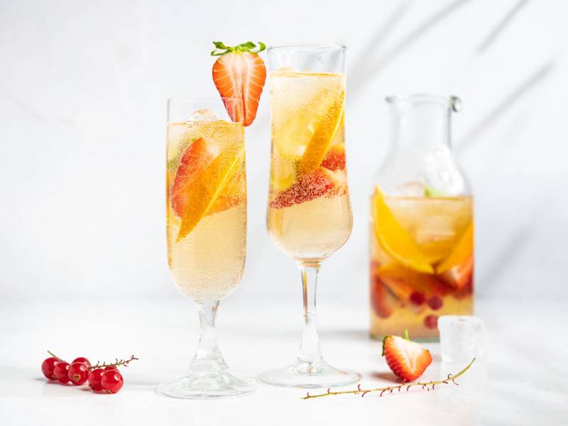 To glass med sommerpunch med frukt og en liten jordbær på kanten, og en mugge med mer sommerpunch i bakgrunnen