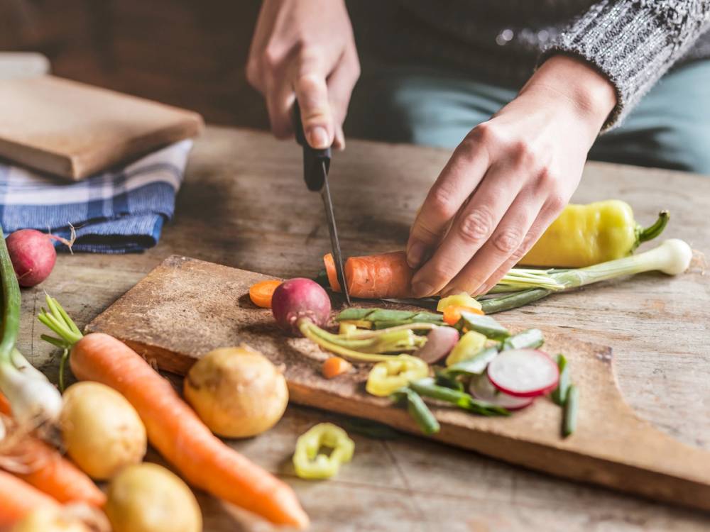 Person skjærer opp gulrøtter og andre grønnsaker på et skjærebrett i et kjøkken.