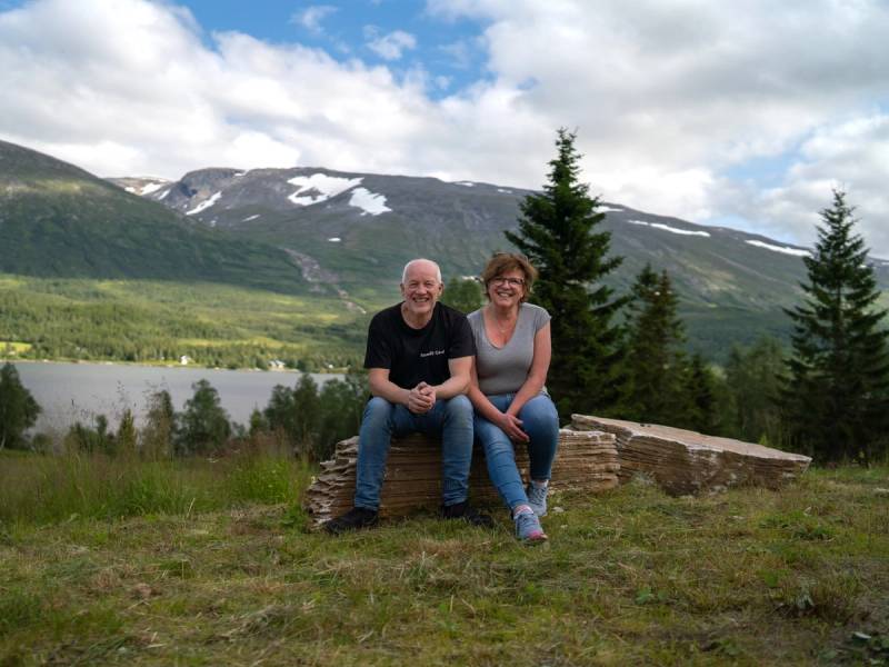 To personer sitter på en stein med fjell, vann og trær i bakgrunnen, under en skyet himmel.