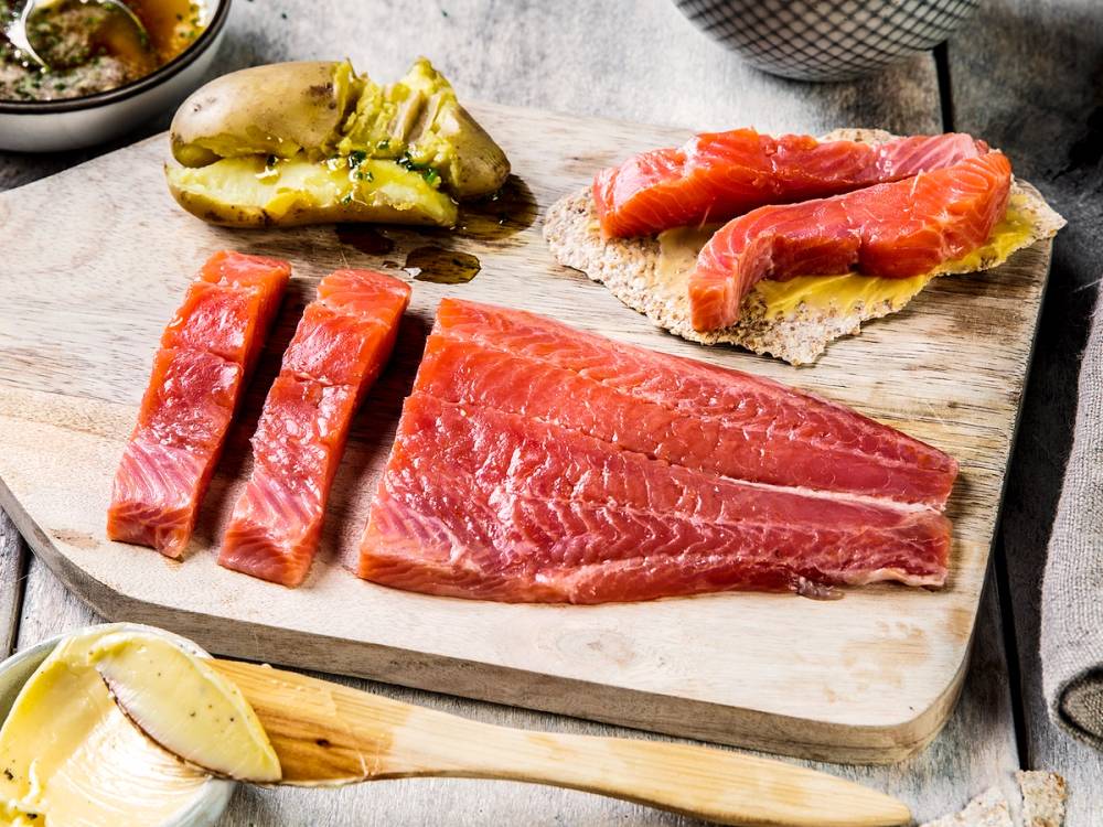 Rakfisk og gravlaks på et skjærebrett med tilbehør som syltet agurk og sitron, klar til servering.