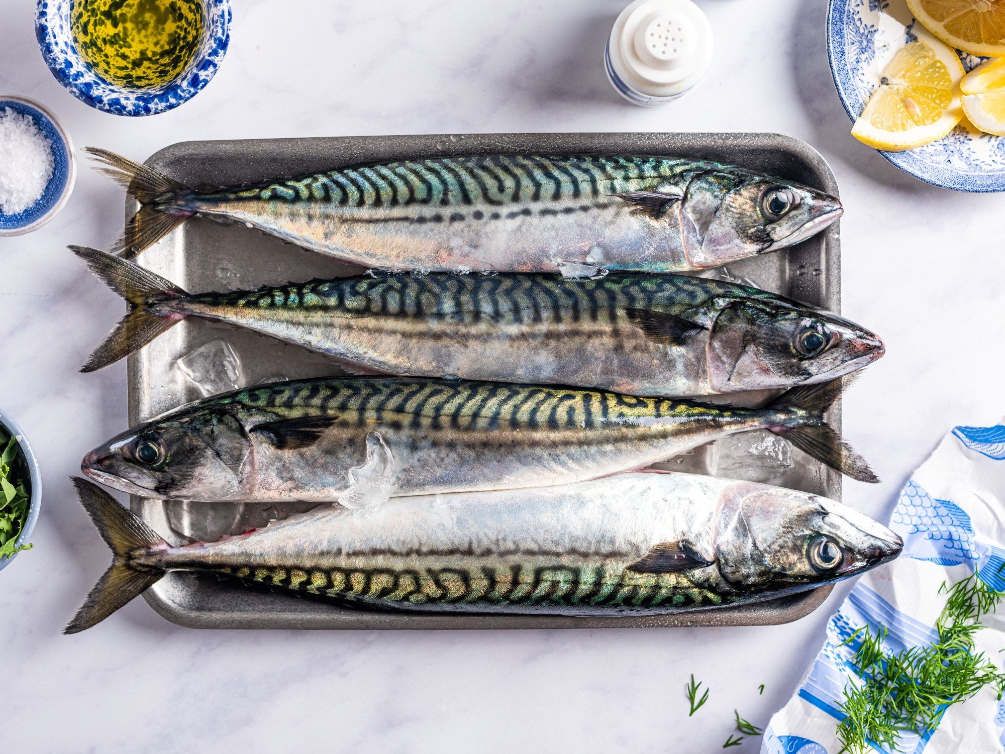 Fire makrellfisker ligger på en metallplate med sitronbåter og urter rundt, klar for tilberedning.