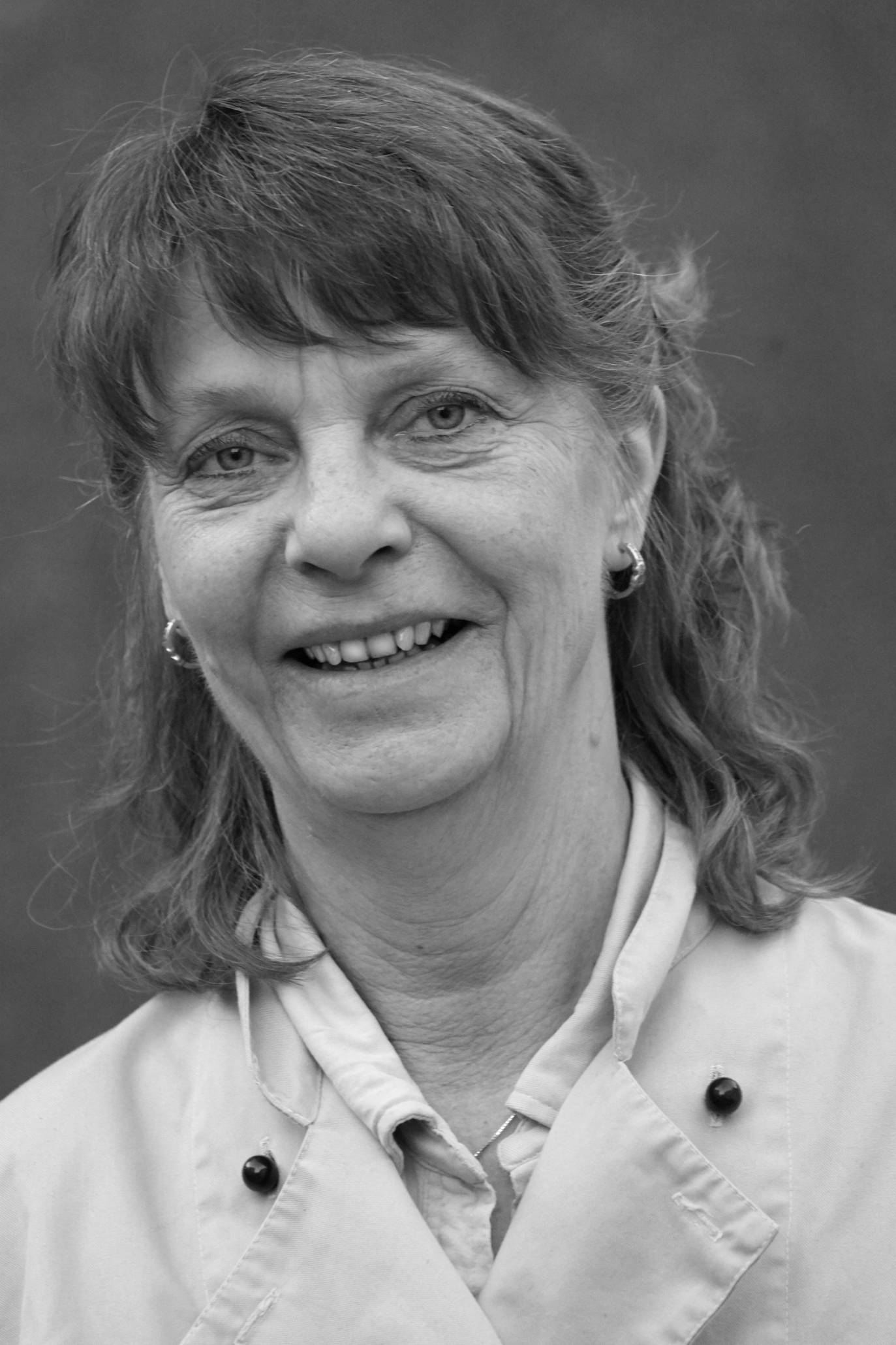 En kvinne med skulderlangt, krøllete hår iført en hvit kokkeforkle, står foran en grå bakgrunn.