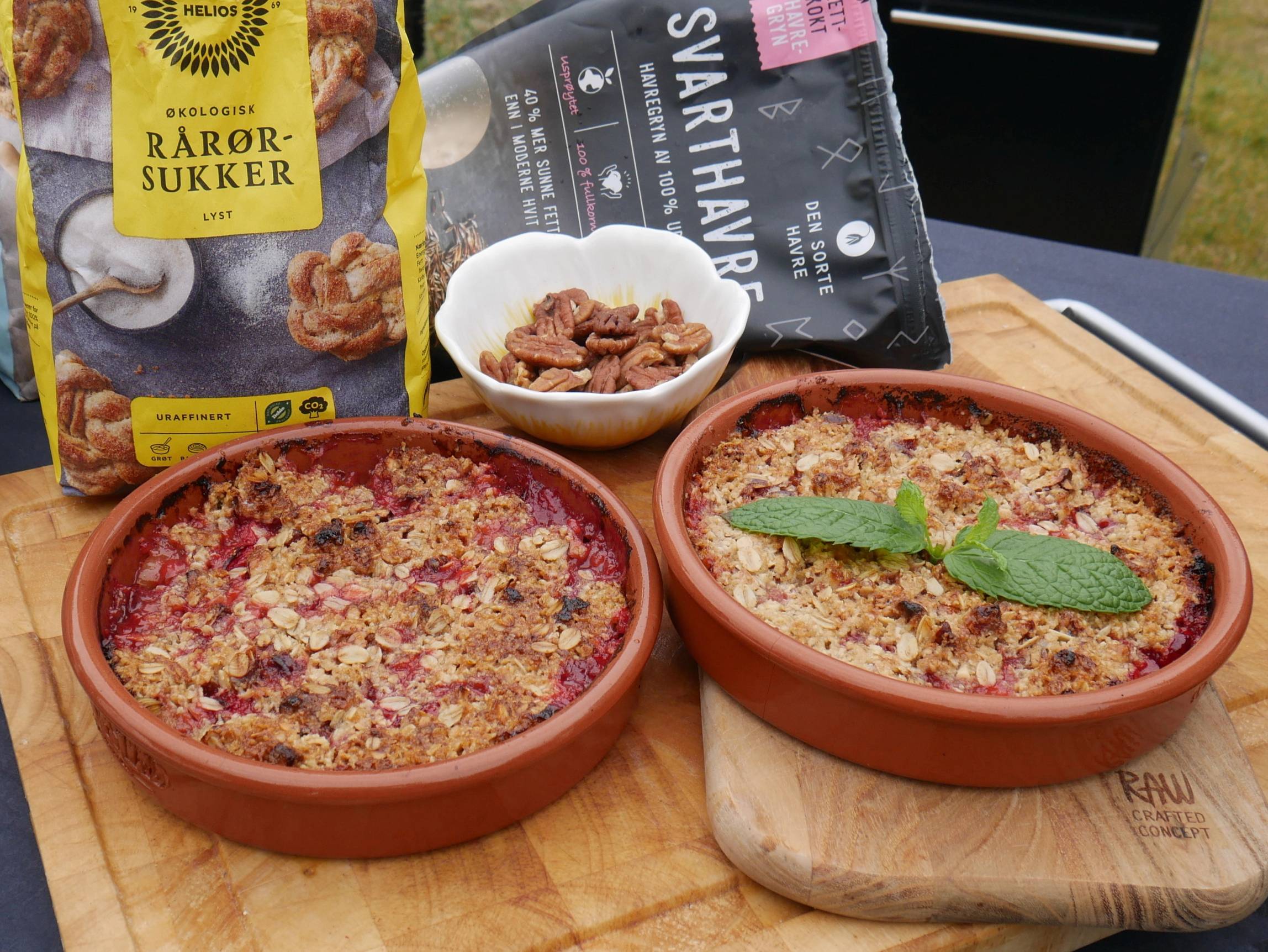 To små ildfaste former med gratinert mat, en med to store salvieblader på toppen, omgitt av en pose med rør-sukker og en pakke med svart pepper, på et trebenkbord i en utendørs setting.