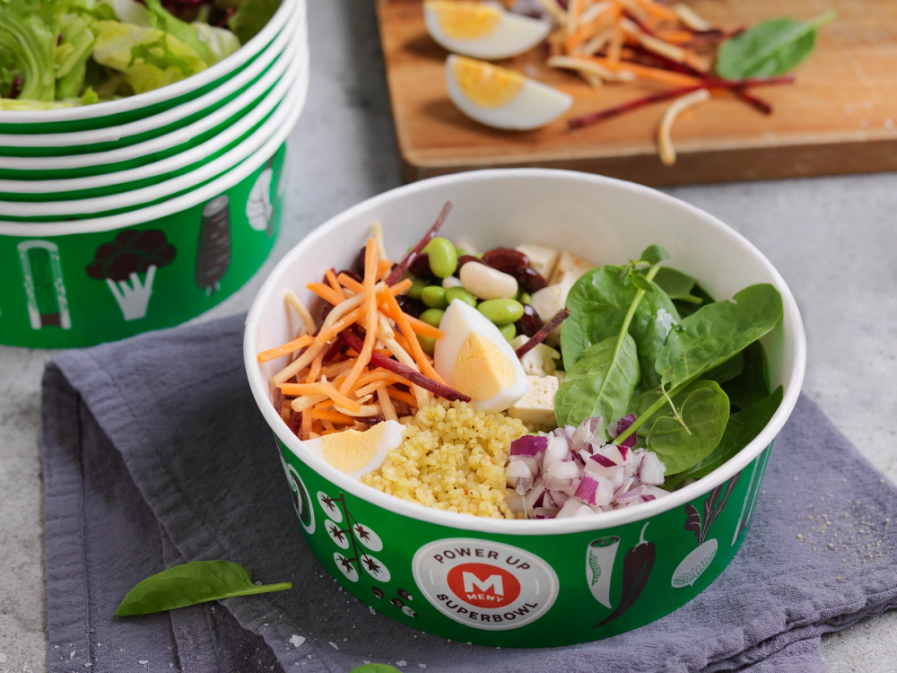 En salat i en grønn salatbar-kopp med ulike grønnsaker og korn, plassert på et bord med en skjærebrett og sitronbåter i bakgrunnen.