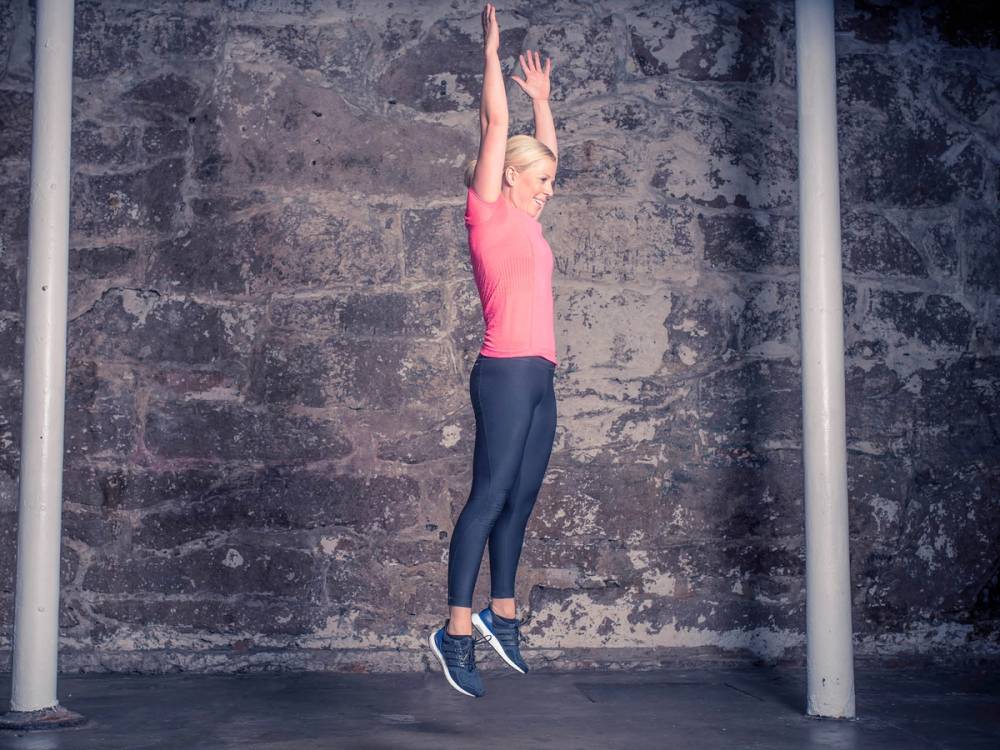 En kvinne gjør burpees med armene hevet over hodet i et treningsrom med en murvegg i bakgrunnen.