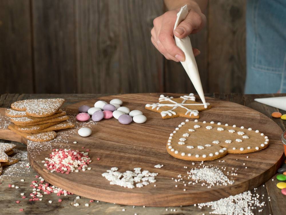 Person pynter en pepperkakehjerte med melis på et rundt trebrett, omgitt av småkaker og sukkerstrøssel.