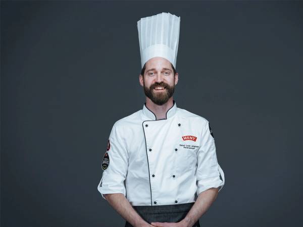 En person kledd som kokk med hvit kokkeuniform og høy kokkehatt, står mot en mørk bakgrunn.