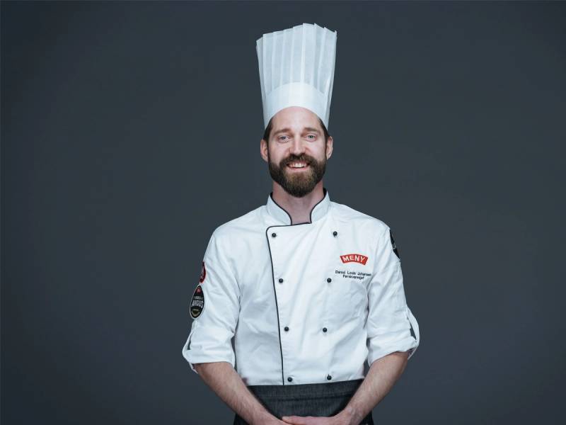 En person kledd som kokk med hvit kokkeuniform og høy kokkehatt, står mot en mørk bakgrunn.