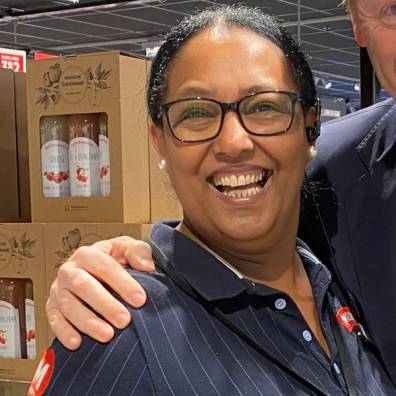 En person i en butikk eller lager med hyller og produkter i bakgrunnen, iført en mørk stripet skjorte og en knapp på brystet.