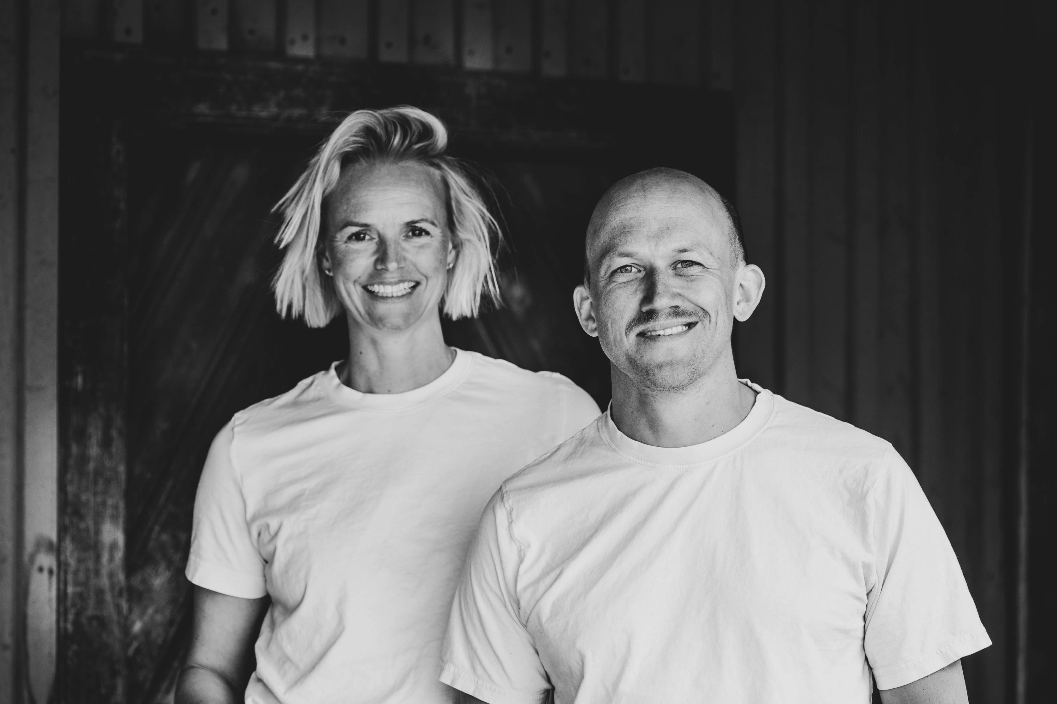 To personer står side om side foran en mørk bakgrunn, begge kledd i hvite t-skjorter, med ansiktene dekket av pixler.