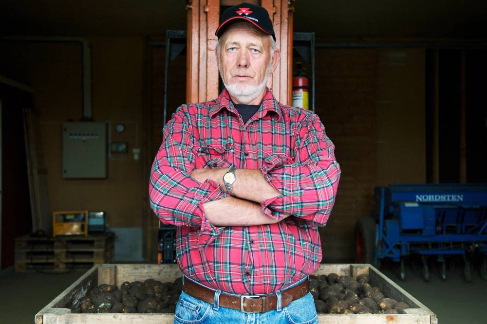 En person står med armene i kors foran en vegg med jord og utstyr, iført rød og blå rutete skjorte og jeans, i en innendørs landbruks- eller lagerlokale.