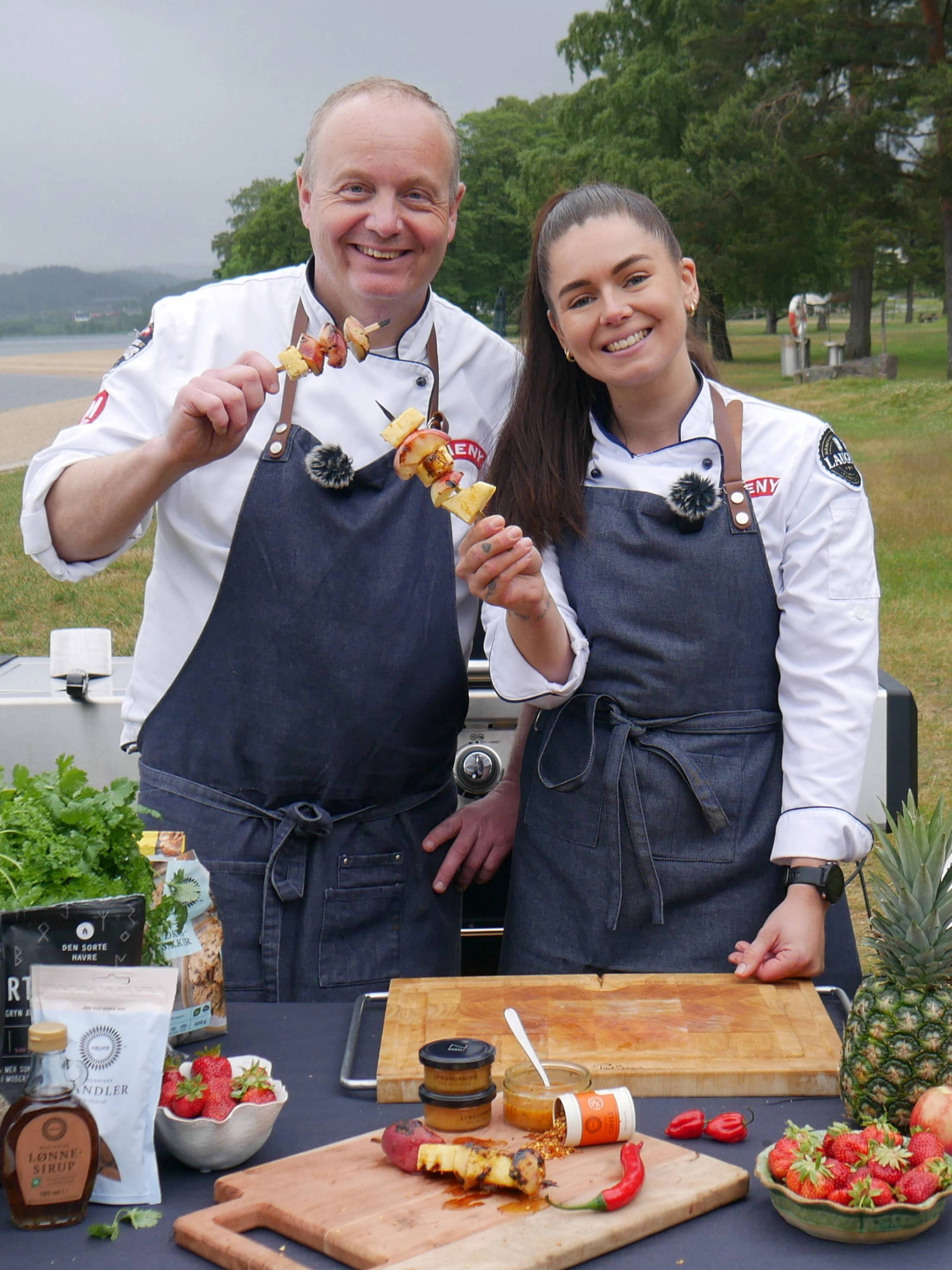 To personer i kokkeuniformer som lager mat utendørs med grilling og frukt på bordet, med vann og trær i bakgrunnen.