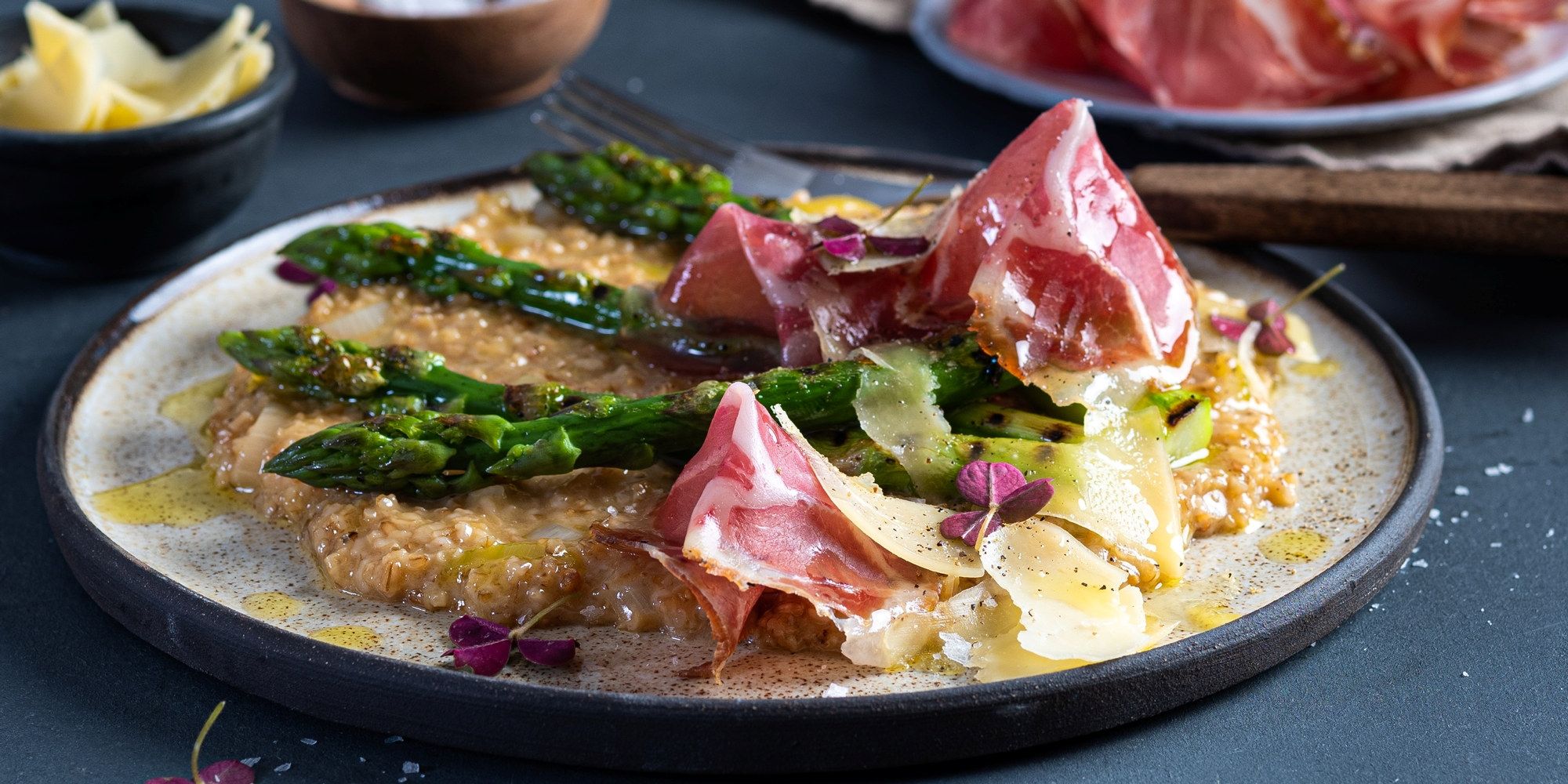 Havreotto med asparges, coppa og ost på en tallerken, servert med grønnsaker og blomsterdekorasjoner, på en mørk bakgrunn.