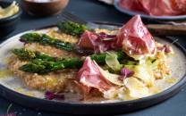 Havreotto med asparges, coppa og ost på en tallerken, servert med grønnsaker og blomsterdekorasjoner, på en mørk bakgrunn.