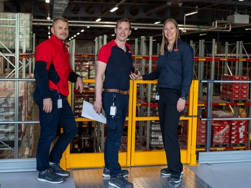 Tre personer i arbeidstøy står i et lager med hyller og varer, og snakker sammen.