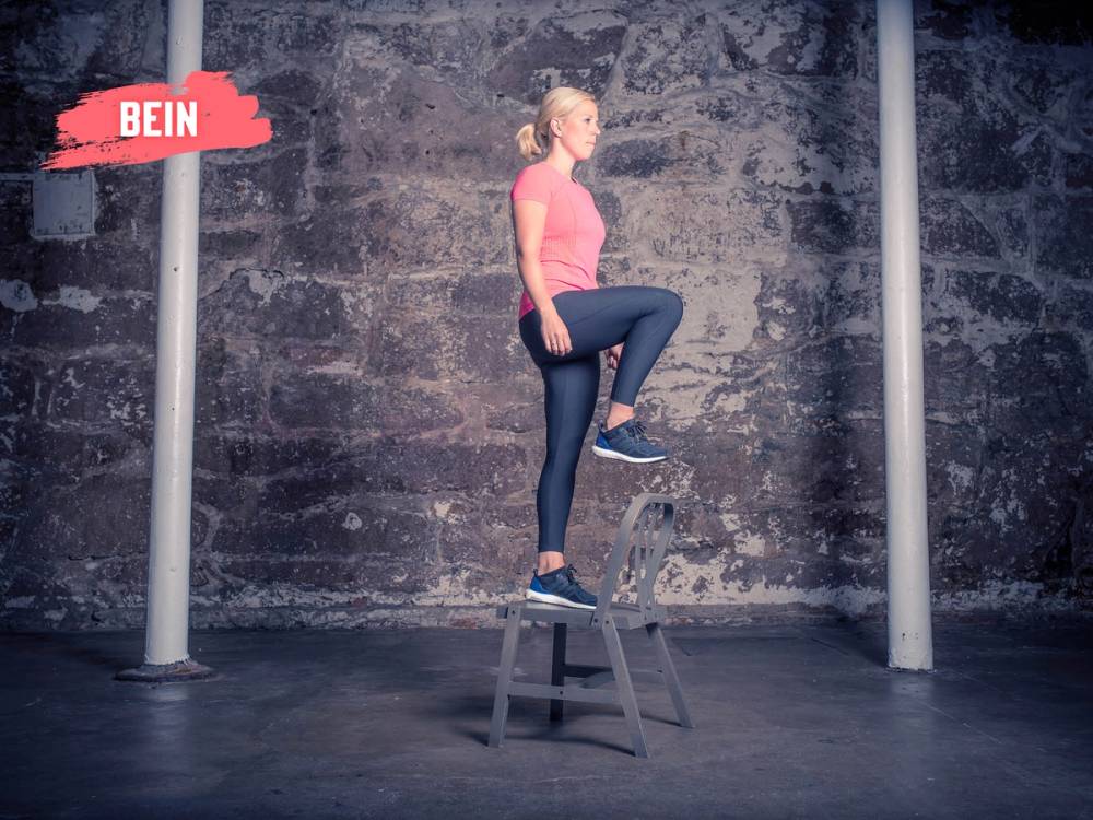 En kvinne står med ett ben på en stol og det andre på en lavere plattform i et treningsrom med en svart vegg i bakgrunnen.