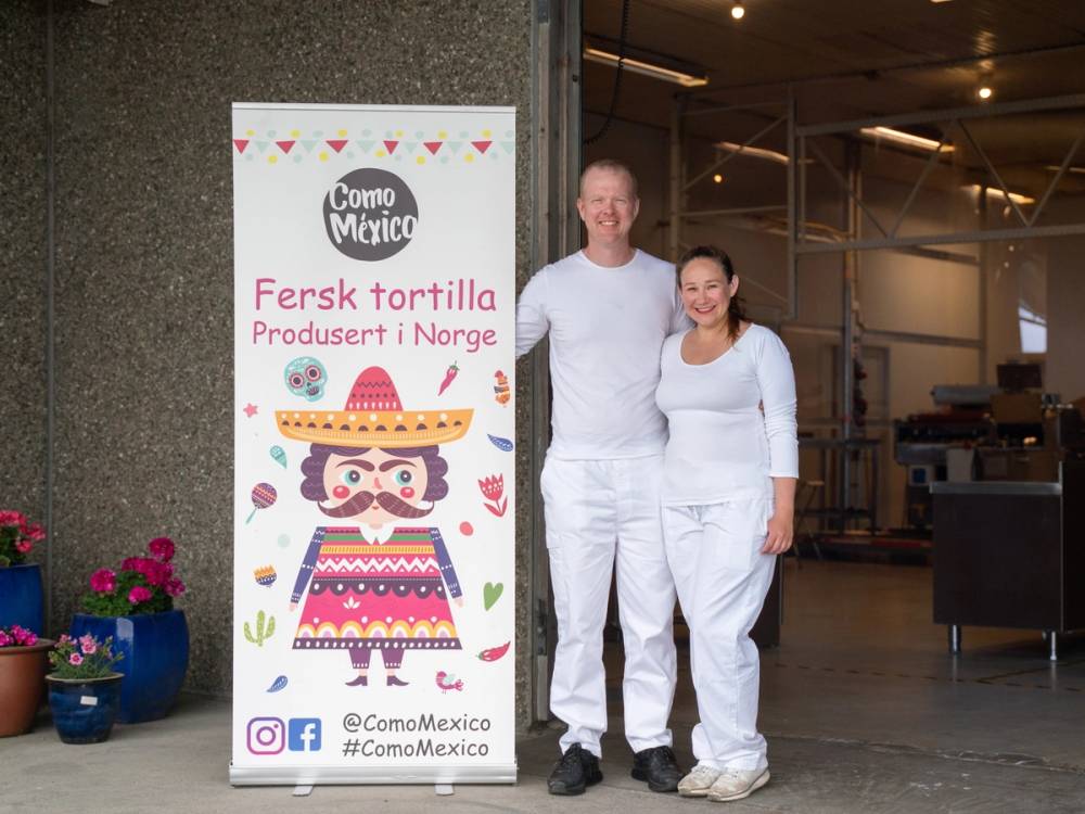 To personer står ved siden av en reklameskilt for autentisk meksikansk mat, med teksten 'Fersk tortilla' og 'Produsert i Norge', foran en butikk eller restaurant. Sikkerhet 90.