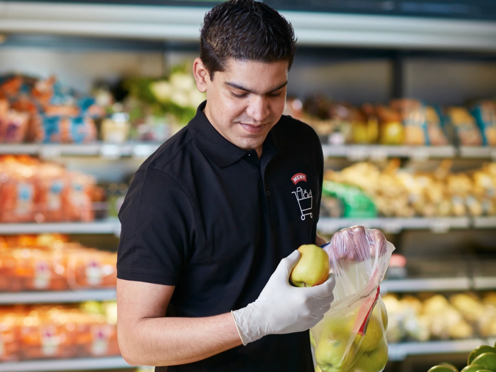 En person i en butikk som plukker frukt, med hyller fylt med ulike varer i bakgrunnen.