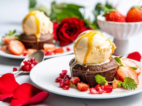 En dessert med sjokoladefondant, vaniljeis og friske bær på en hvit tallerken, omgitt av roseblader og en kopp med jordbær i bakgrunnen.