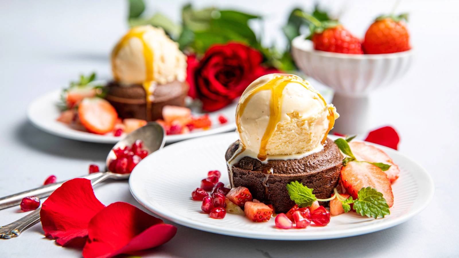 En dessert med sjokoladefondant, vaniljeis og friske bær på en hvit tallerken, omgitt av roseblader og en kopp med jordbær i bakgrunnen.