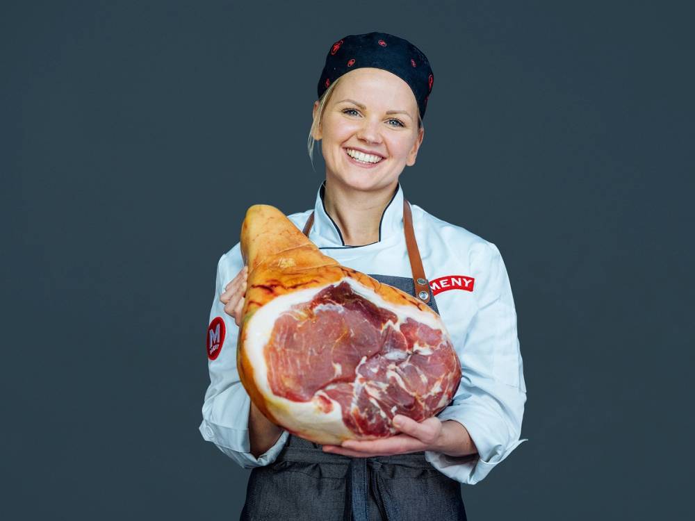 En person i arbeidsklær holder en stor skinke og en skinkebit i hendene, med en mørk bakgrunn.