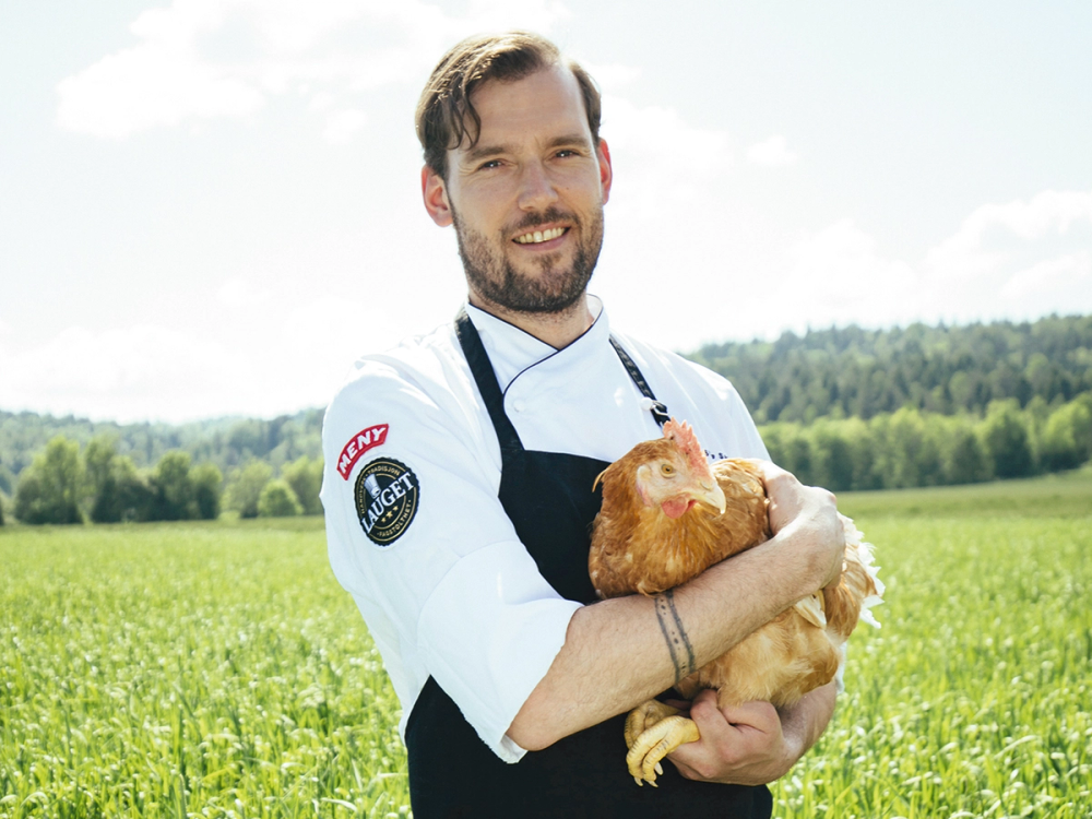 En person i kokkeforkle holder en kylling i armene ute på en grønn eng med trær i bakgrunnen.