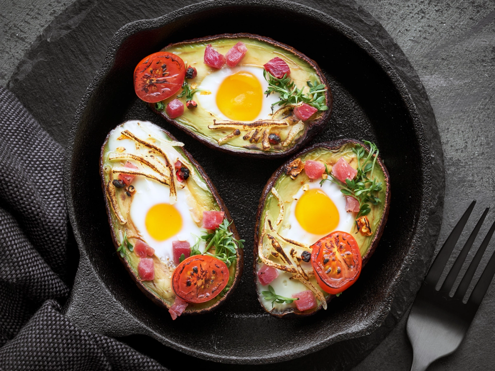 Tre halverte avokado fylt med skinke og egg, pyntet med cherrytomater og urter, på en svart tallerken.