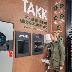 Kvinne bruker avfalls- og glassautomater ved en miljøstasjon, med en stor takk-tekst på veggen som takker for riktig avfallshåndtering.