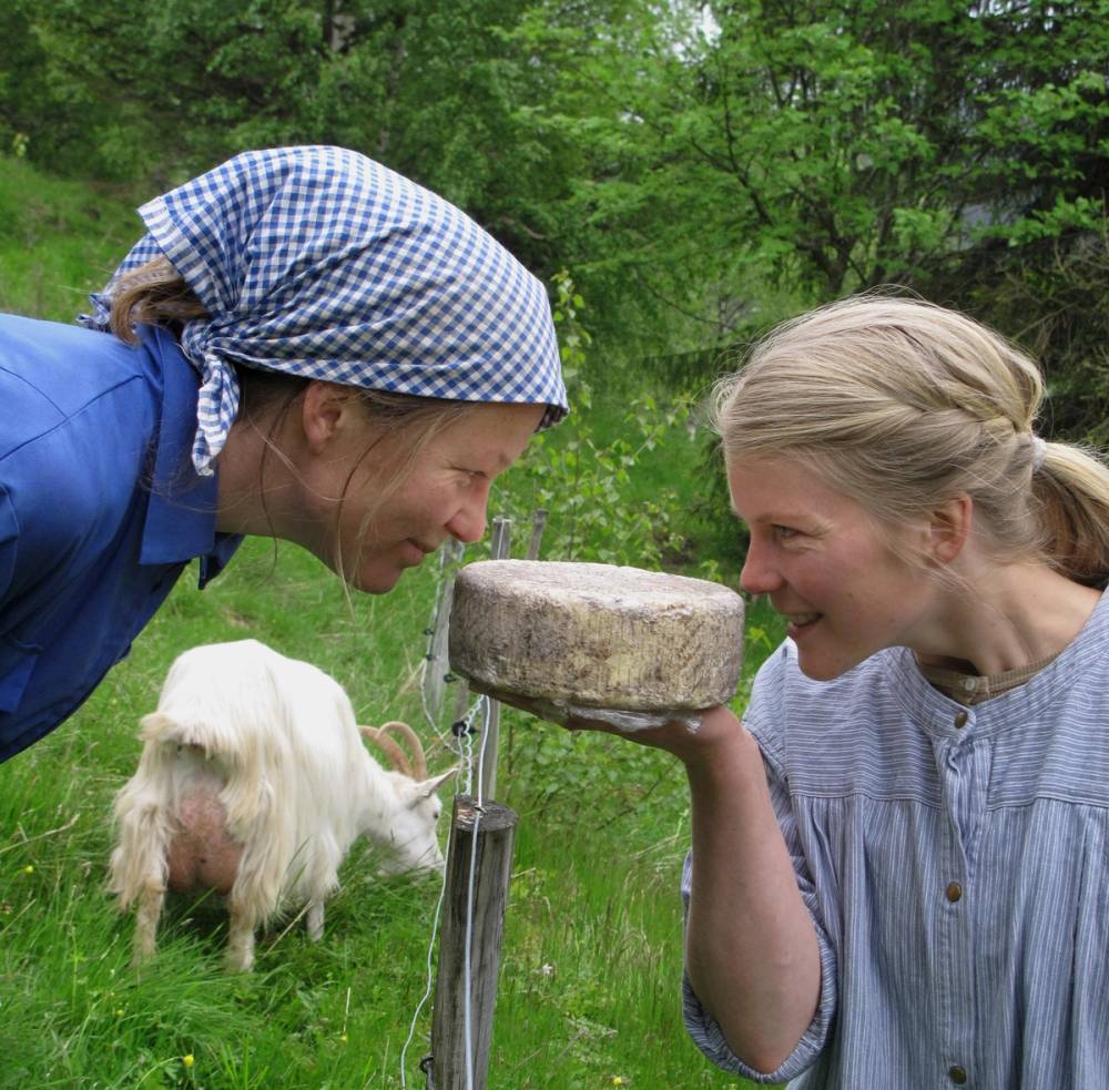 To personer som drikker fra en stor ost på en utendørs gresslette med en liten hund i bakgrunnen, omgitt av grønne trær.