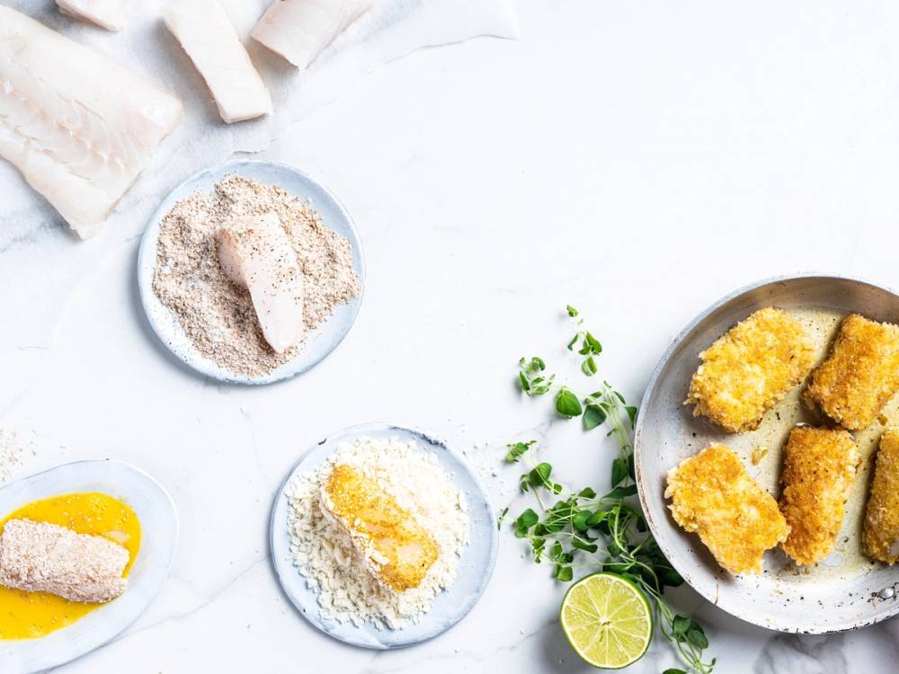 Steg-for-steg bilde av fiskepinner med tilbehør som sitron, krydder og brødsmuler på små tallerkener og en skål med fiskepinner på en hvit bakgrunn.