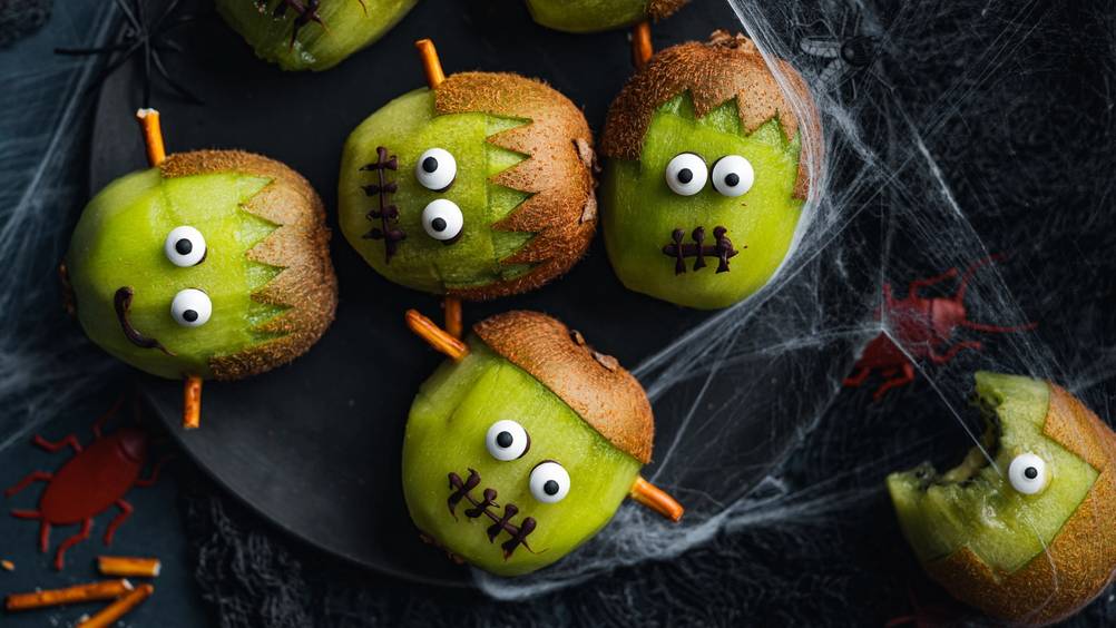 Eplepyntede snacks med ansikter og små horn, dekorert til Halloween, på en svart tallerken med spindelvev rundt.