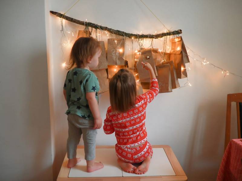 To barn lager en julekalender på en vegg med lys og dekorasjoner, en av barna peker mot kalenderen.