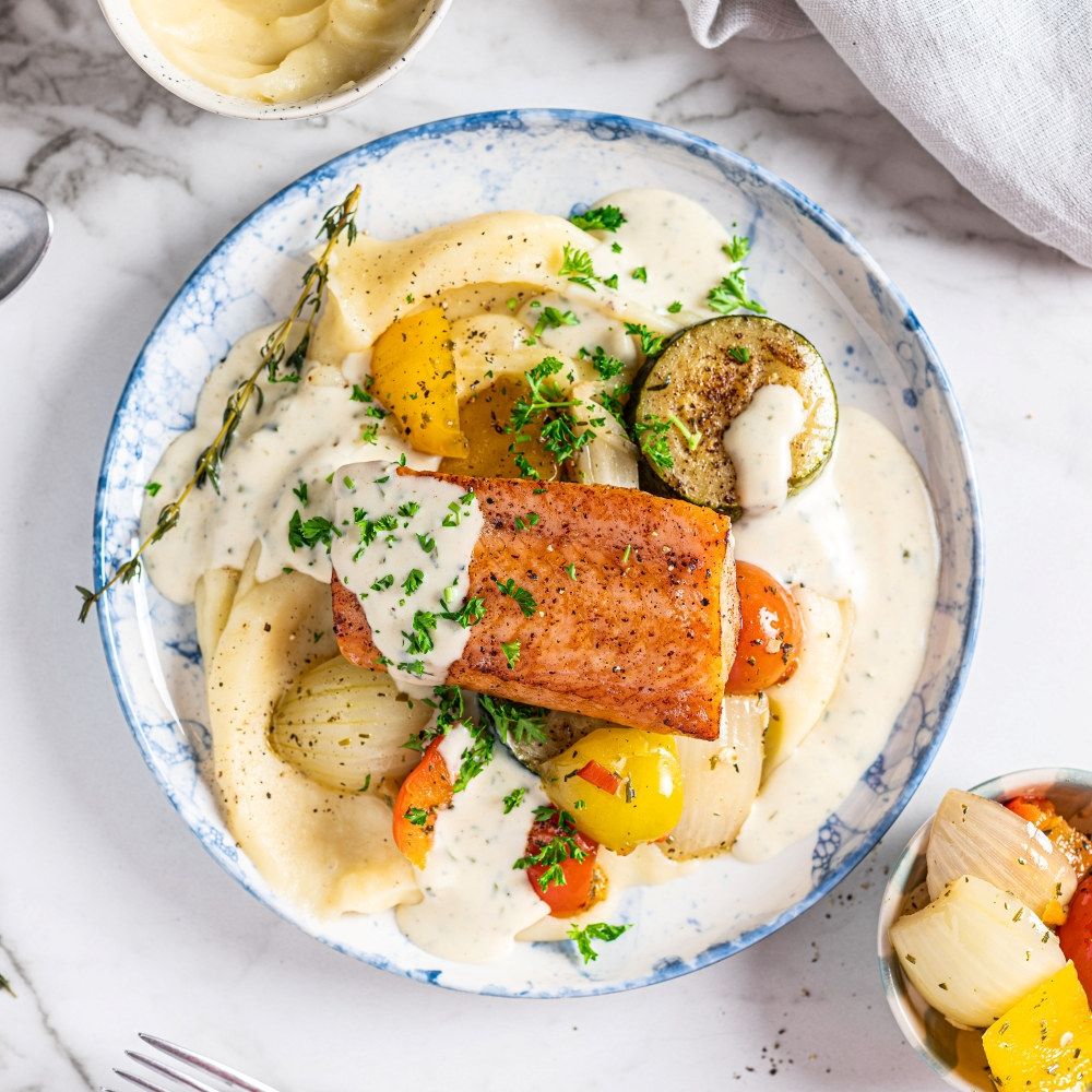 Laks med potetmos og grønnsaker på en hvit tallerken med saus og ferske urter.