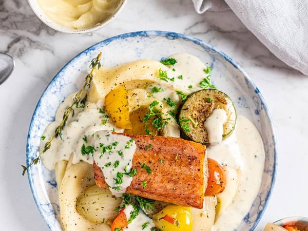 Laks med potetmos og grønnsaker på en hvit tallerken med saus og ferske urter.