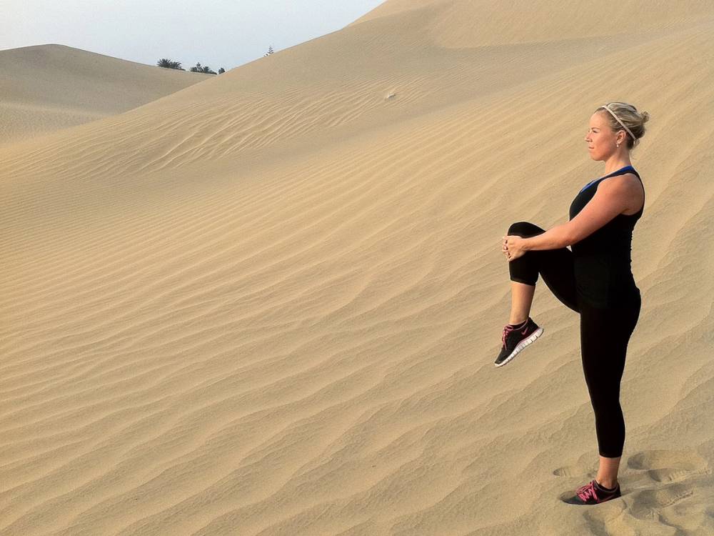 En person står på sanddyner i ørkenen og ser ut over landskapet, med armene rundt knærne.