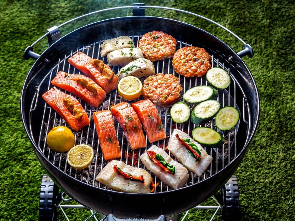 Fisk og grønnsaker grilles på en utendørs grill med gressbakgrunn.