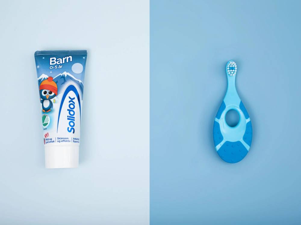 En tube med barnetannkrem og en blå tannbørste med hvite prikker ligger på en lys blå bakgrunn.