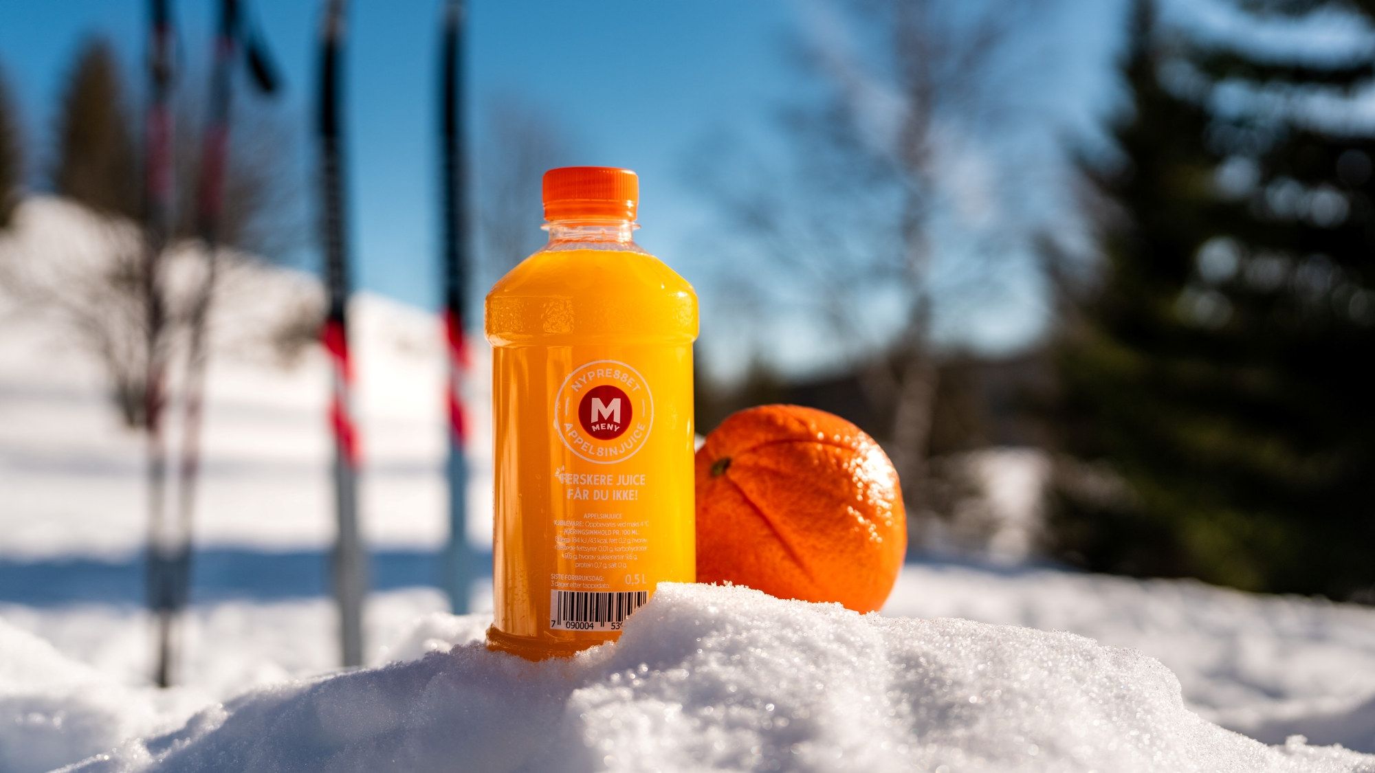 En flaske med gul drikke og en appelsin ligger på snø med ski og trær i bakgrunnen