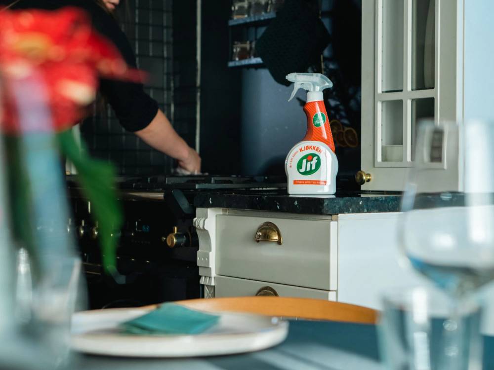 En person vasker kjøkkenbenken med en sprayflaske, mens andre personer er i bakgrunnen.