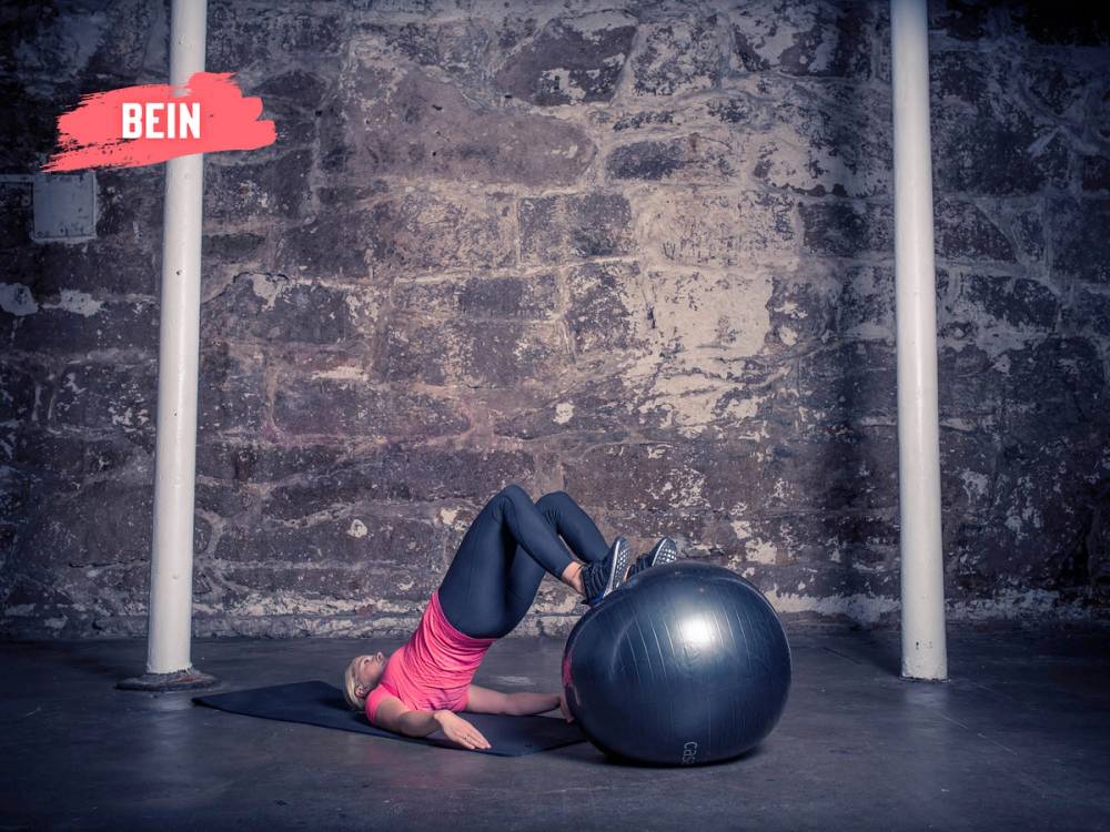 En person ligger på ryggen med beina bøyd og føttene på en treningsball, mens de trener på gulvet i et treningsrom.